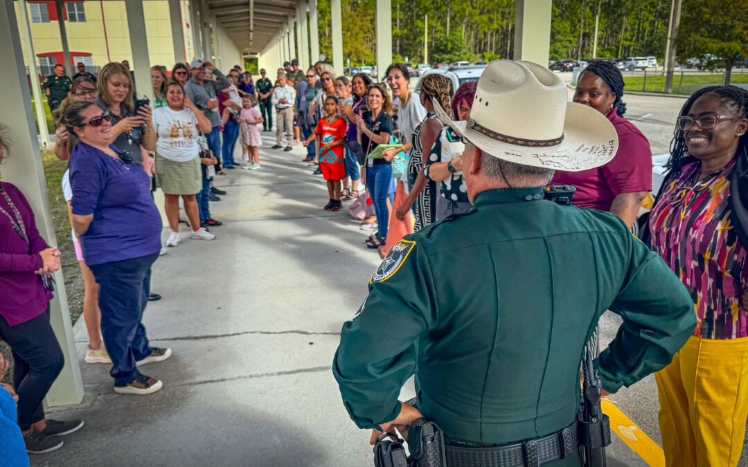 A police officer in uniform, seen from behind, stands facing a long line of smiling and engaged adults and children outside a building on a covered walkway The group appears to be participating in a welcoming or celebratory event