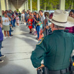 A police officer in uniform, seen from behind, stands facing a long line of smiling and engaged adults and children outside a building on a covered walkway The group appears to be participating in a welcoming or celebratory event