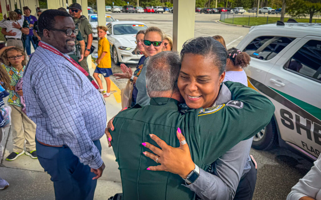 A smiling woman in a sheriff’s uniform hugs a male officer outside near a sheriff’s vehicle, while onlookers, including children and adults, observe the joyful moment