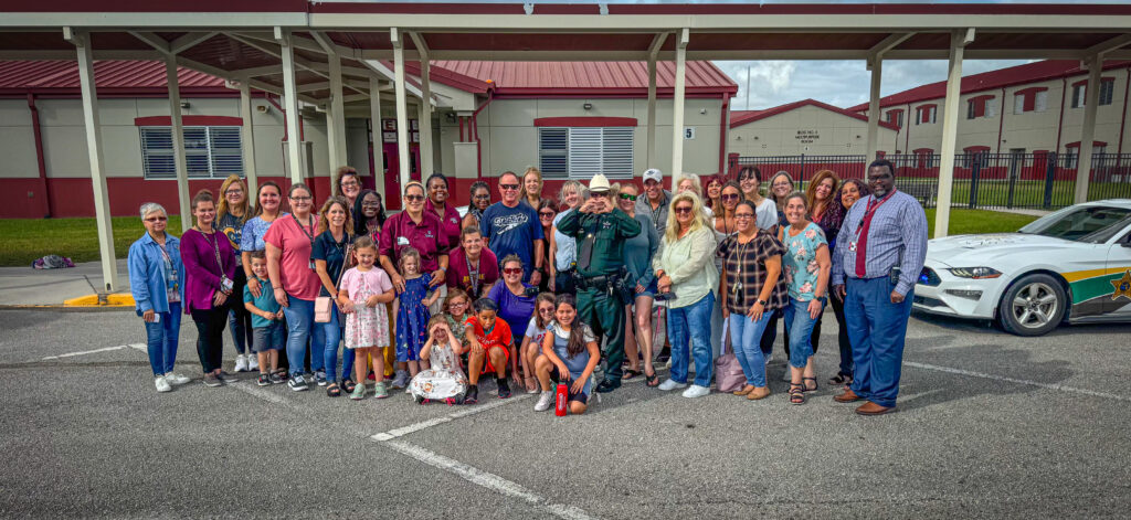 A large group of adults and children pose and smile together in a parking lot outside a school building A police officer stands in the middle, and a patrol car is parked to the right