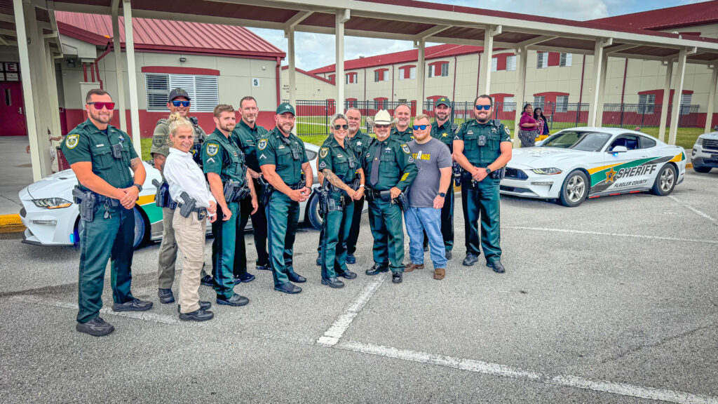 A group of uniformed sheriff’s deputies and two civilians stand smiling in front of two sheriff patrol cars in a parking lot outside a building with red roofs