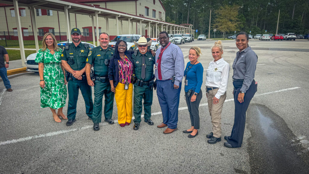 A group of people, including law enforcement officers in uniform and individuals in business and casual attire, stand together smiling in a parking lot in front of a large building