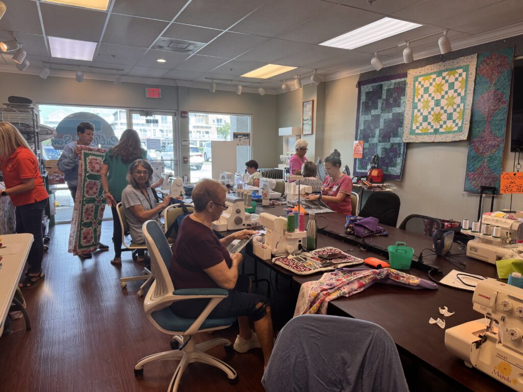 A group of women work on sewing machines and quilts in a well lit craft room Finished quilts hang on the wall, and some people stand and chat while others sew at tables Supplies and fabrics are scattered around