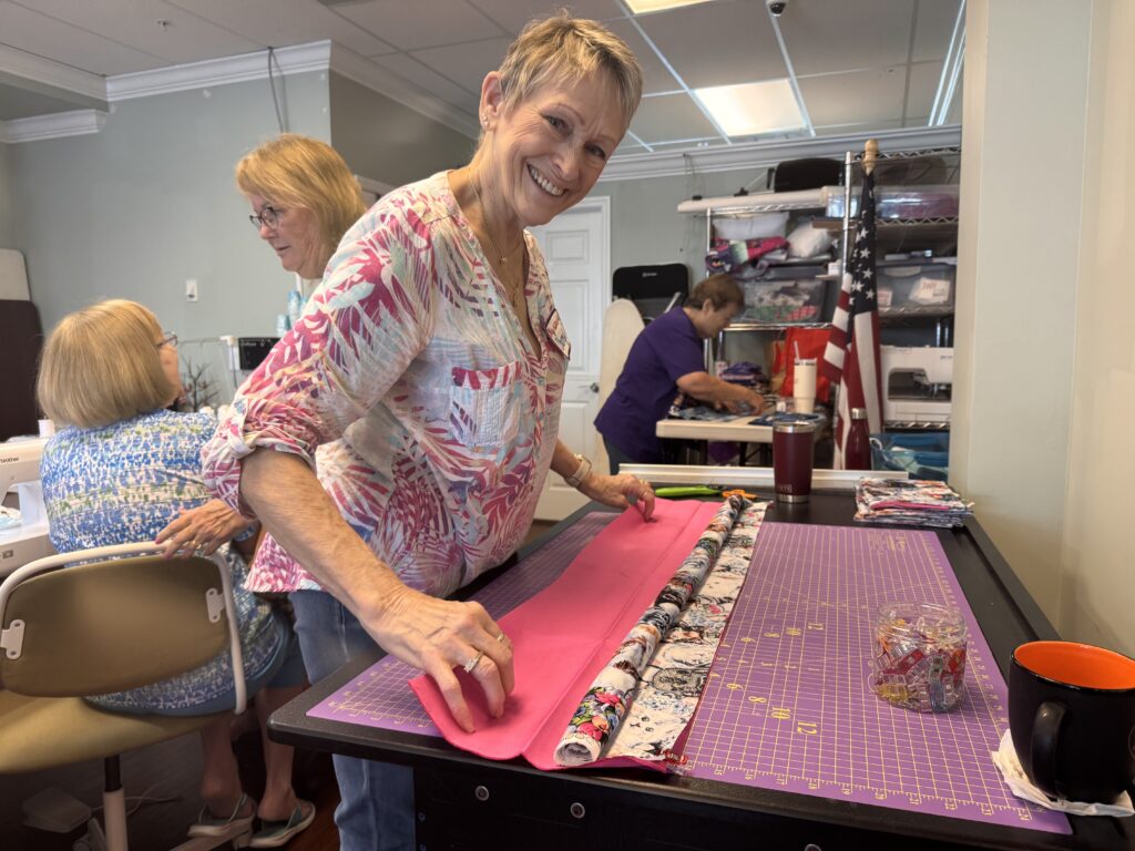 A smiling woman stands at a table, folding pink and patterned fabric Three other women work at sewing machines in a craft room filled with supplies A mug and sewing tools sit on the table