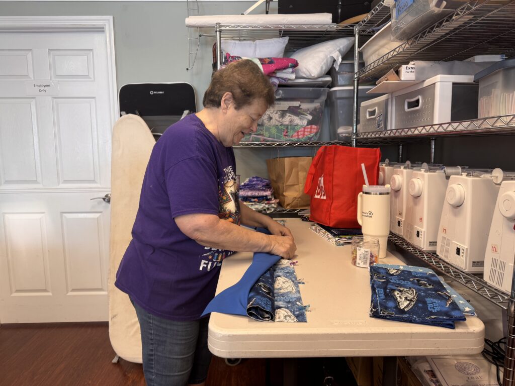 A woman in a purple shirt stands at a table, pinning or cutting blue fabric Sewing machines, fabric, and storage containers are organized on metal shelves beside her in a craft room