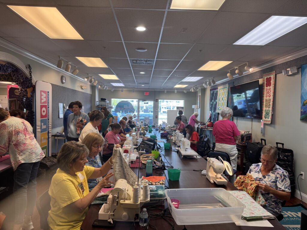 A large group of women are gathered in a brightly lit room, sitting at long tables and working on sewing projects with sewing machines, fabric, and craft supplies Quilts and colorful fabrics decorate the walls