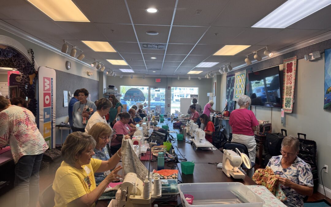 A large group of women are gathered in a brightly lit room, sitting at long tables and working on sewing projects with sewing machines, fabric, and craft supplies Quilts and colorful fabrics decorate the walls