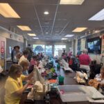 A large group of women are gathered in a brightly lit room, sitting at long tables and working on sewing projects with sewing machines, fabric, and craft supplies Quilts and colorful fabrics decorate the walls