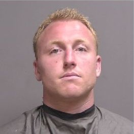 A man with short blond hair and a serious expression is shown against a plain background, wearing a dark cape like garment, in a mugshot style photo
