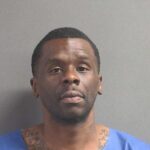A man with short hair and visible neck tattoos wearing a blue garment stands against a plain gray background, looking slightly above the camera—reminiscent of booking photos from Flagler County