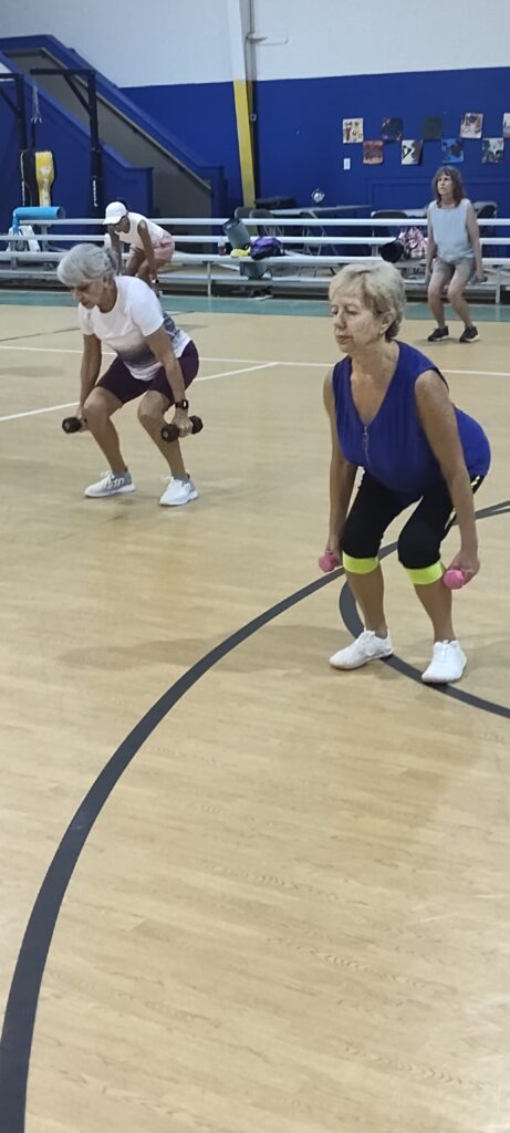 A group of older adults exercise in a gym, performing squats while holding small dumbbells They are dressed in athletic wear and appear focused and active Benches and gym equipment are visible in the background
