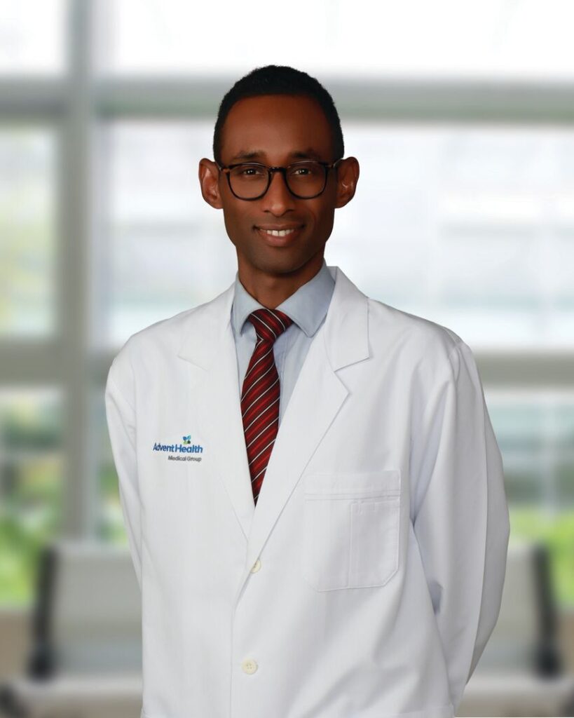 A man wearing glasses, a white lab coat with an "Advent Health" logo, and a red striped tie stands indoors in a bright, modern medical facility