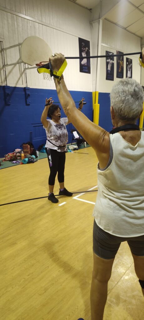 Two older women exercise in a gymnasium, one in the foreground raising her arms, and another facing her and mirroring the movement Gym bags and exercise mats are visible in the background