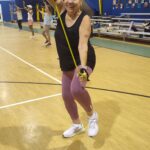 An older woman in athletic wear smiles while exercising with resistance bands in a gym Other people are working out in the background The gym has high ceilings and wooden floors
