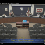 A wide view of a county commission meeting room shows officials seated at a curved desk, with U S and Florida flags behind them The room is sparsely attended, and the screen displays workshop details and the date //
