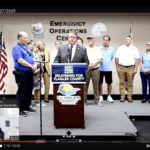 A man speaks at a podium labeled "Delivering for Flagler County" during a press conference at the Emergency Operations Center Several people stand behind him, with U S and state flags displayed to the left