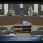A county commissioners meeting is in session with officials seated at a curved desk, audience members in front, and presentation screens visible U S and Florida flags are displayed The date "//" appears on screen