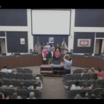 A group of people stands at the front of a school board meeting room while an audience sits and someone takes a photo of the group American and Florida flags are displayed behind them