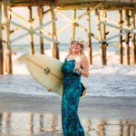 A smiling woman in a blue patterned dress and cowboy hat stands on the beach holding a surfboard and a trophy, with a wooden pier and ocean waves in the background