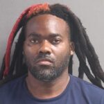 A man with long dreadlocks, some dyed red, wearing a navy blue shirt, poses against a neutral gray background in Flagler County