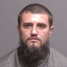 A man with short brown hair and a full beard looks directly at the camera He is wearing a gray, high collared shirt, and the background is plain and neutral