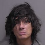 A person with long, messy dark hair and a serious expression looks directly at the camera against a plain background