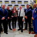Six people in formal attire stand smiling together at a Crime Stoppers event The woman in the center holds a plaque The background features Crime Stoppers banners and balloon decorations