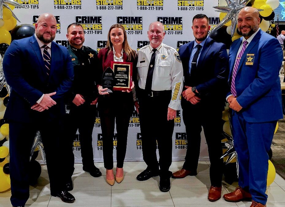 Six people in formal attire stand smiling together at a Crime Stoppers event The woman in the center holds a plaque The background features Crime Stoppers banners and balloon decorations