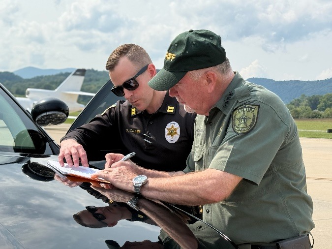 Two law enforcement officers review documents on the hood of a black car at an airport, with an airplane and mountains visible in the background on a sunny day