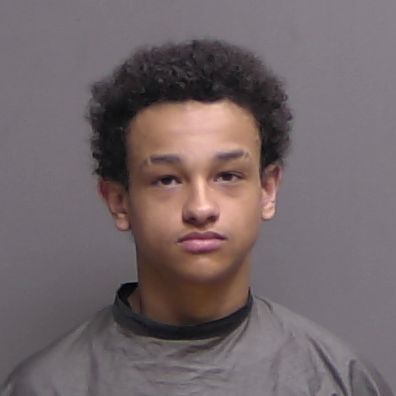 A young person with short curly hair, wearing a dark colored garment, stands against a plain, neutral background and looks directly at the camera with a neutral expression A young person with short curly hair, wearing a dark colored garment, stands against a plain, neutral background and looks directly at the camera with a neutral expression