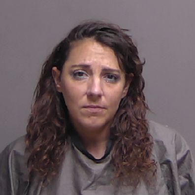 A woman with wavy brown hair wearing a gray garment stands against a plain, neutral background, looking directly at the camera with a serious expression A woman with wavy brown hair wearing a gray garment stands against a plain, neutral background, looking directly at the camera with a serious expression