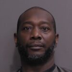 A middle aged man with short hair and a trimmed beard, wearing a dark shirt, faces the camera against a plain gray background