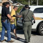 Two law enforcement officers escort a man in a yellow shirt and flip flops toward a sheriff’s vehicle parked in a residential driveway The man’s hands appear restrained behind his back