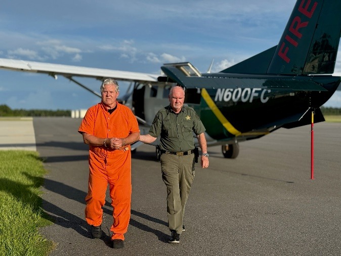 A man in an orange jumpsuit with handcuffs is escorted by a uniformed officer across an airstrip, with a small airplane marked "NFC" in the background A man in an orange jumpsuit with handcuffs is escorted by a uniformed officer across an airstrip, with a small airplane marked "NFC" in the background