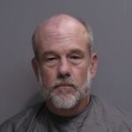 A middle aged man with a bald head and gray beard is wearing a gray shirt and facing the camera against a plain gray background