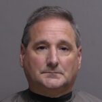 A middle aged man with short gray hair wearing a dark colored shirt is shown against a plain gray background, looking directly at the camera with a neutral expression A middle aged man with short gray hair wearing a dark colored shirt is shown against a plain gray background, looking directly at the camera with a neutral expression