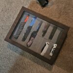 A wooden display case with a glass top, containing ten folding pocket knives of various colors and designs, is placed on a beige carpet A small black object lies nearby on the carpet