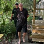 A barefoot man in a black shirt and shorts is being escorted by a uniformed police officer along a path outside a building surrounded by greenery