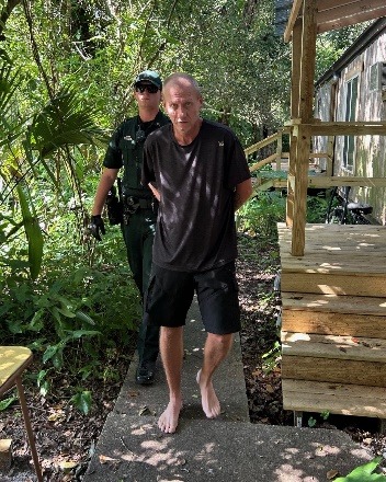 A barefoot man in a black shirt and shorts is being escorted by a uniformed police officer along a path outside a building surrounded by greenery