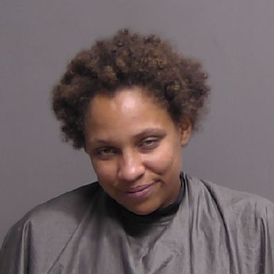 A person with short curly hair wearing a gray drape is standing against a plain gray background, looking slightly downward with a subtle smile A person with short curly hair wearing a gray drape is standing against a plain gray background, looking slightly downward with a subtle smile