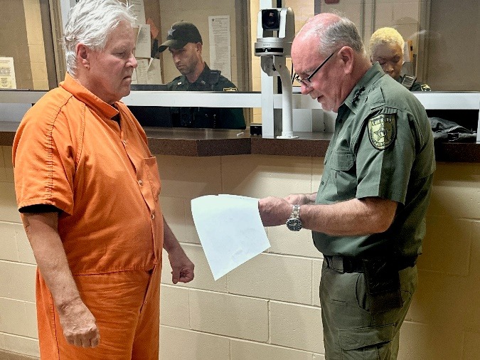 A man in an orange prison jumpsuit stands facing a uniformed officer who is reading a document Two more officers are visible in the background behind a counter The setting appears to be a jail or detention facility