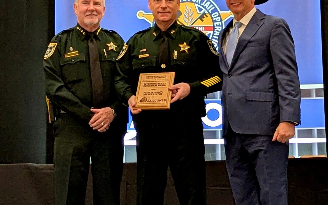 Three men stand together at an awards event Two are in sheriff uniforms and one is in a suit and cowboy hat The man in the center holds a plaque A blue banner with a sheriff’s badge logo is in the background
