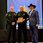 Three men stand together at an awards event Two are in sheriff uniforms and one is in a suit and cowboy hat The man in the center holds a plaque A blue banner with a sheriff’s badge logo is in the background