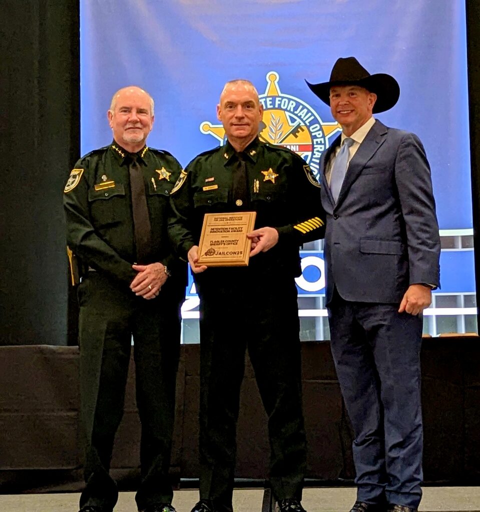 Three men stand together at an awards event Two are in sheriff uniforms and one is in a suit and cowboy hat The man in the center holds a plaque A blue banner with a sheriff’s badge logo is in the background