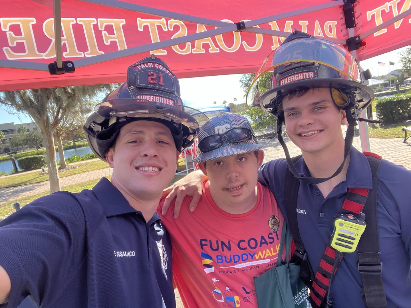 Three smiling people pose under a red tent labeled "COAST FIRE " Two wear firefighter helmets and navy uniforms, and the person in the middle wears a red Buddy Walk T shirt and baseball cap Outdoor setting with trees and blue sky