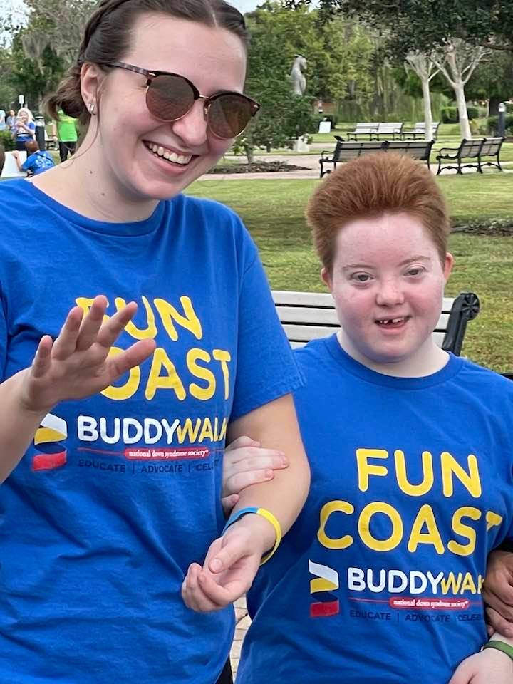 Two people wearing matching blue "Fun Coast Buddy Walk" shirts stand arm in arm outdoors, smiling One person waves while the other stands close, with trees and benches visible in the background