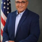 A man in a blue suit and glasses poses in front of an American flag, smiling slightly with his hands resting on a table The background is plain and gray