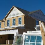 A two story house under construction with exposed wooden framing, partially installed windows, and building materials stacked in front of it, set against a clear blue sky