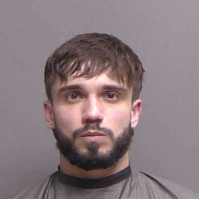 A young man with short brown hair and a full beard looks directly at the camera against a plain gray background, wearing a high collared, dark garment A young man with short brown hair and a full beard looks directly at the camera against a plain gray background, wearing a high collared, dark garment