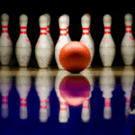 A bright orange bowling ball rolls toward a set of seven white bowling pins with red stripes, reflected on the shiny blue surface of the bowling lane